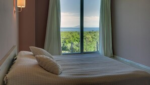 Une chambre à coucher avec un grand lit, deux oreillers, une table de chevet, une lampe fixée au mur et une fenêtre avec des rideaux.