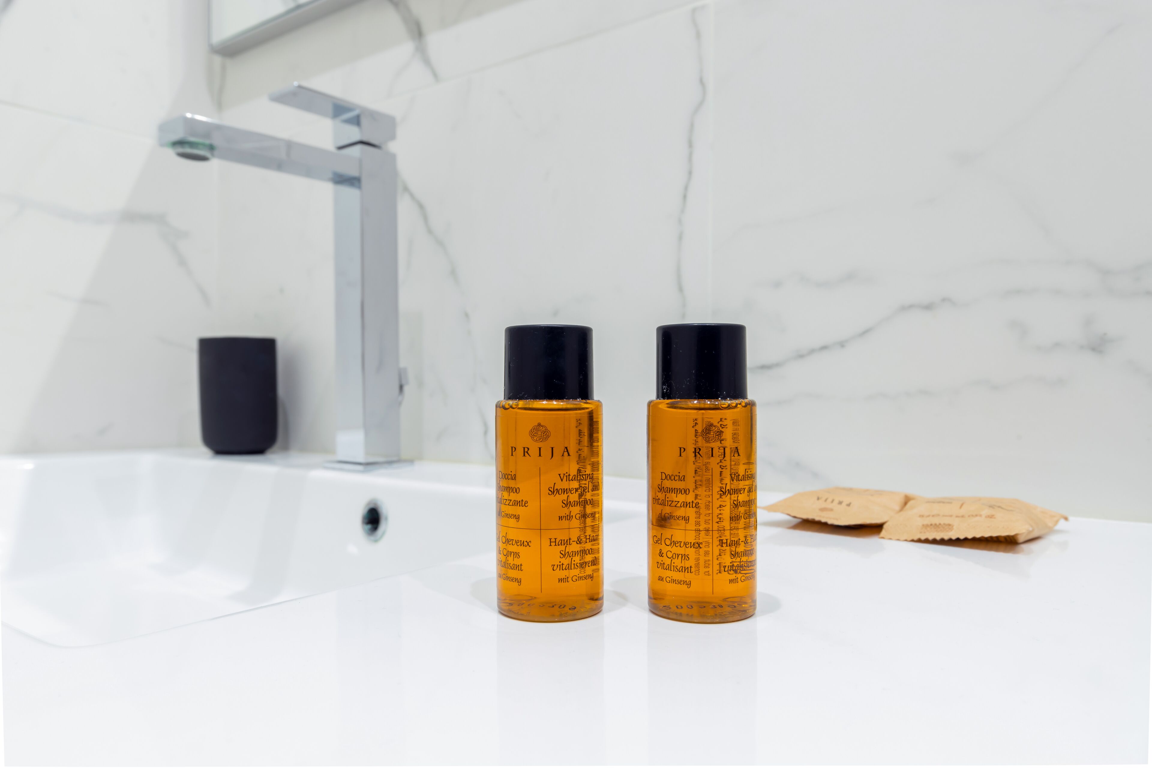 Two bottles of PRIJIA shampoo and conditioner on a white bathroom sink with a marble backsplash.