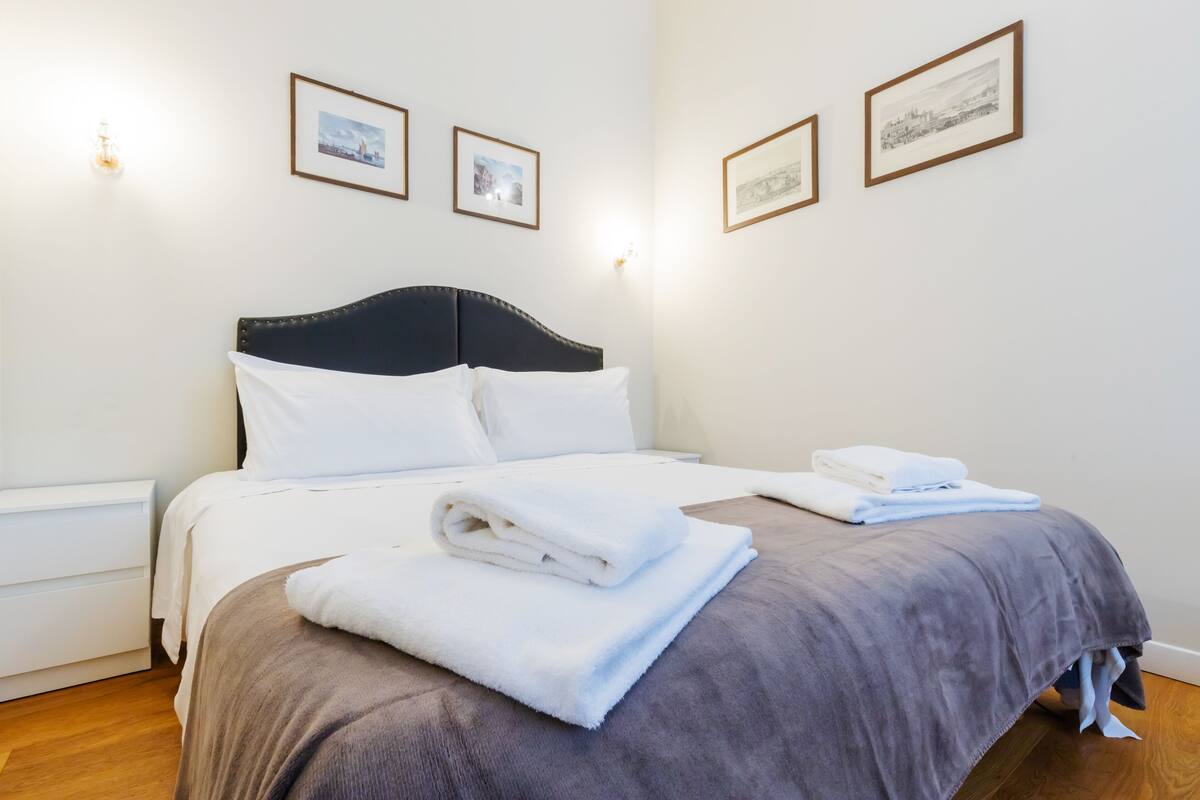 A neatly made bed with white and grey linens, a wooden nightstand, and framed pictures on the wall.
