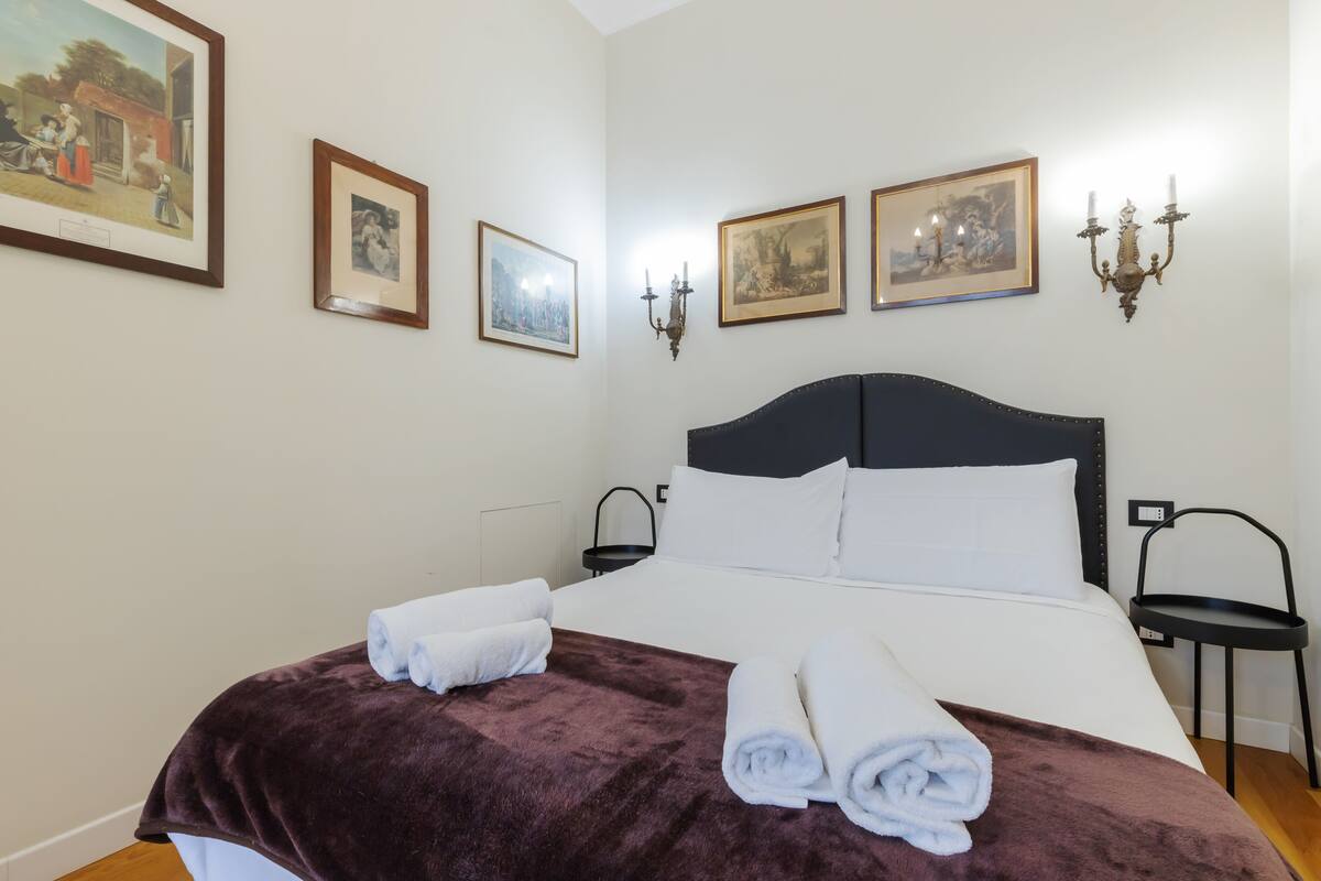 A neatly made bed with white and brown linens, a headboard, and a dark headboard. There are two wall-mounted sconces and three framed pictures on the wall.