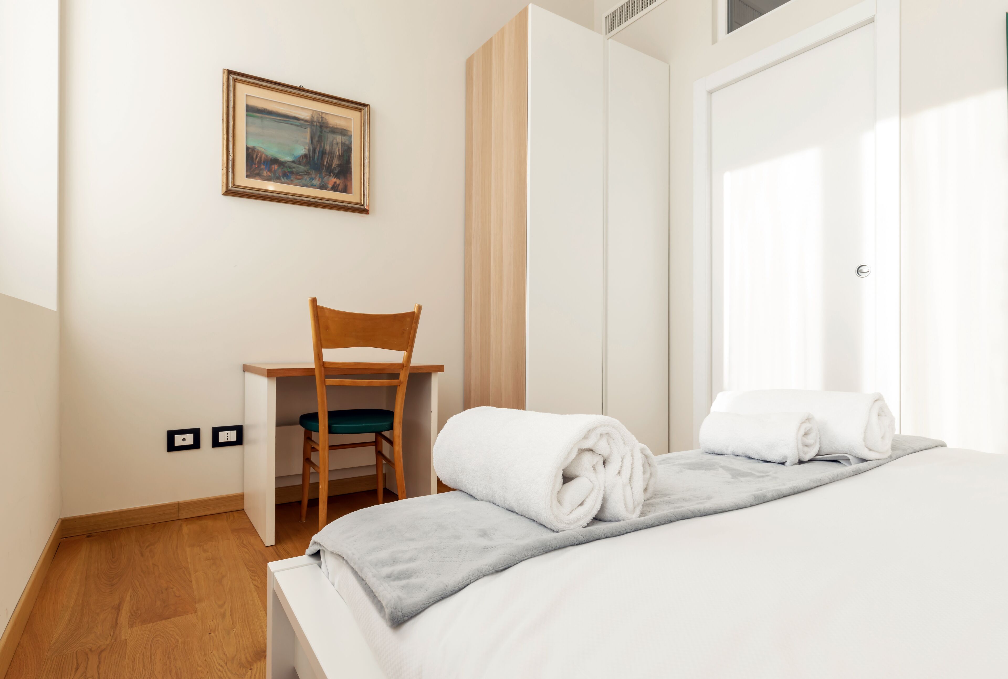 A hotel room with a bed, a desk, a chair, and a painting on the wall.