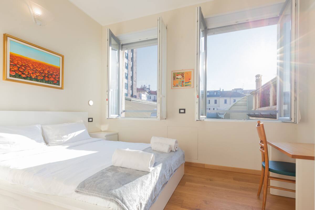 A bedroom with a bed, two windows, a desk, and a chair.