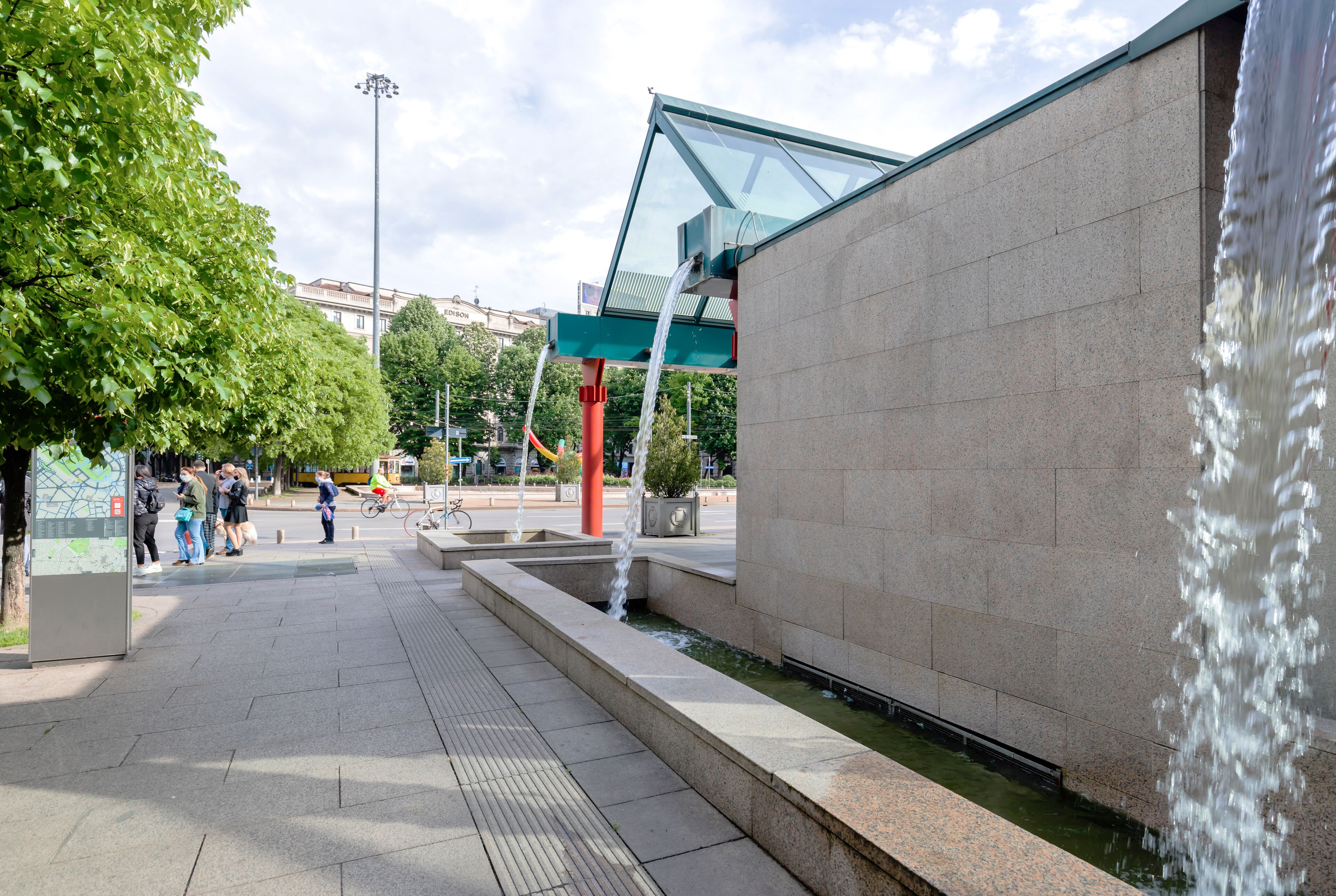 Ein öffentlicher Platz mit Wasserspiel, einer Wegweiser-Karte und einem modernen Gebäude mit Glasdach.