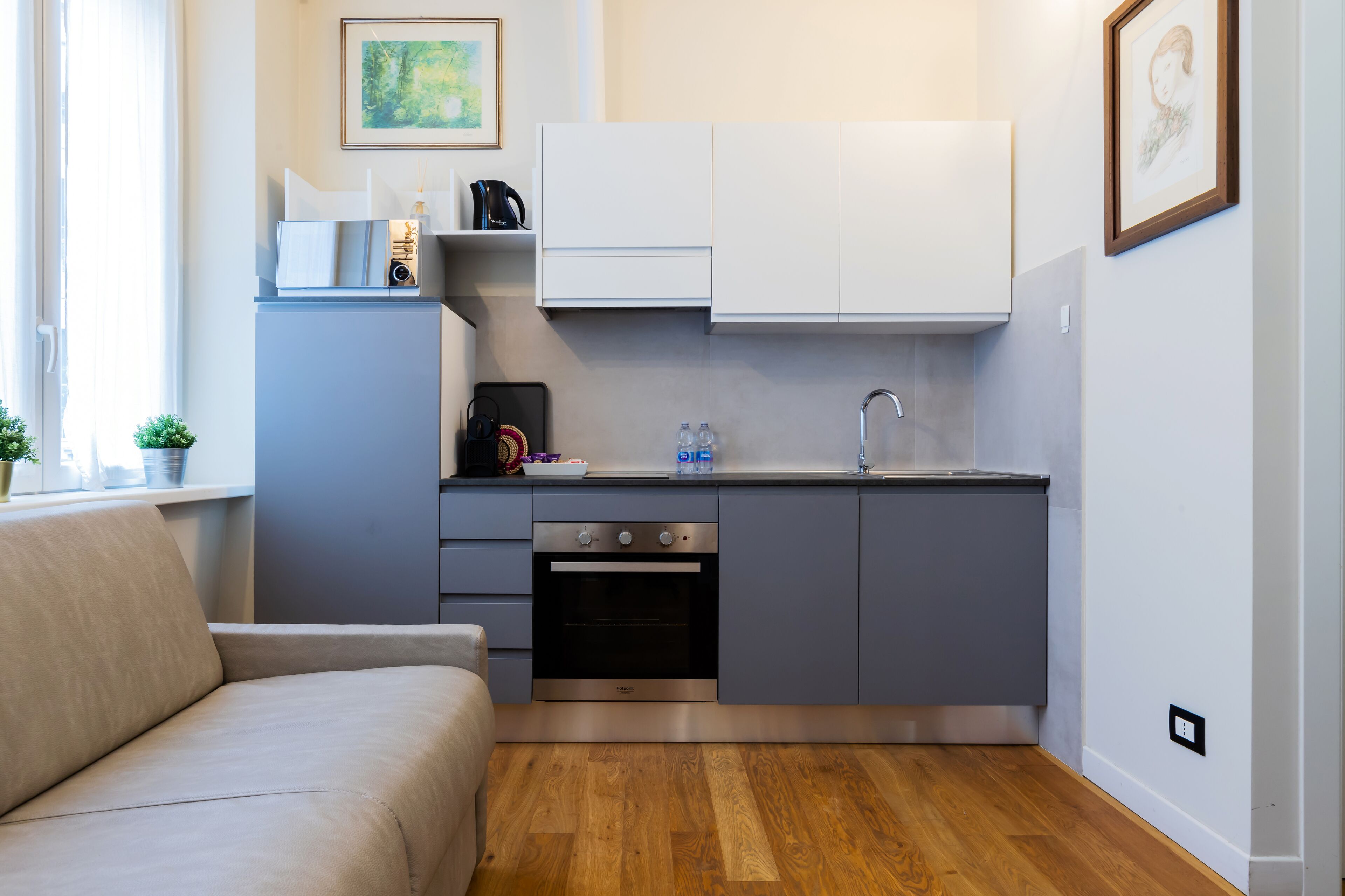 A compact kitchen with grey cabinets, a built-in oven, and a sink. There is a small countertop with a kettle and a framed picture above it.