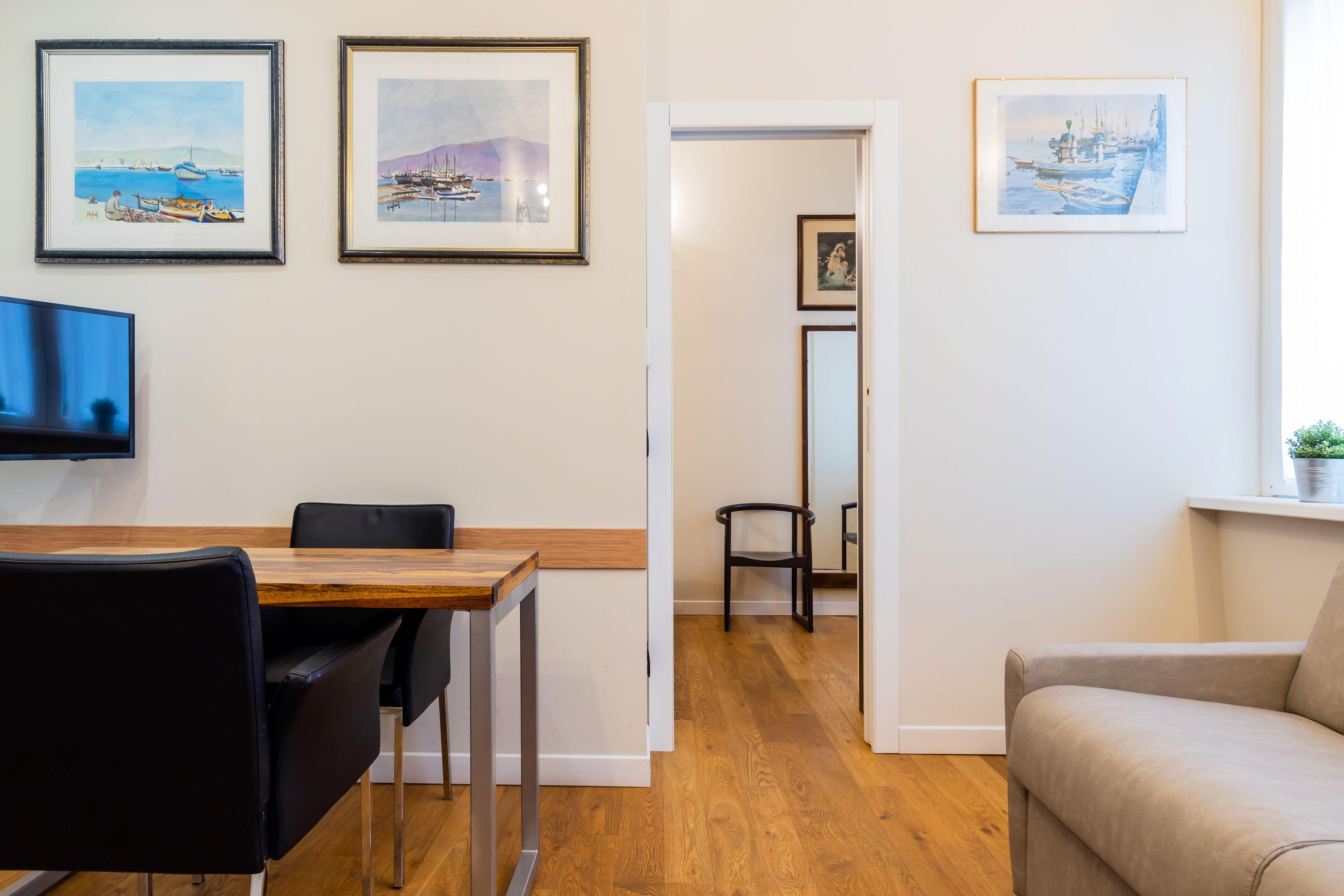 A modern living room with a wooden dining table, black chairs, a beige sofa, and framed artwork on the walls.