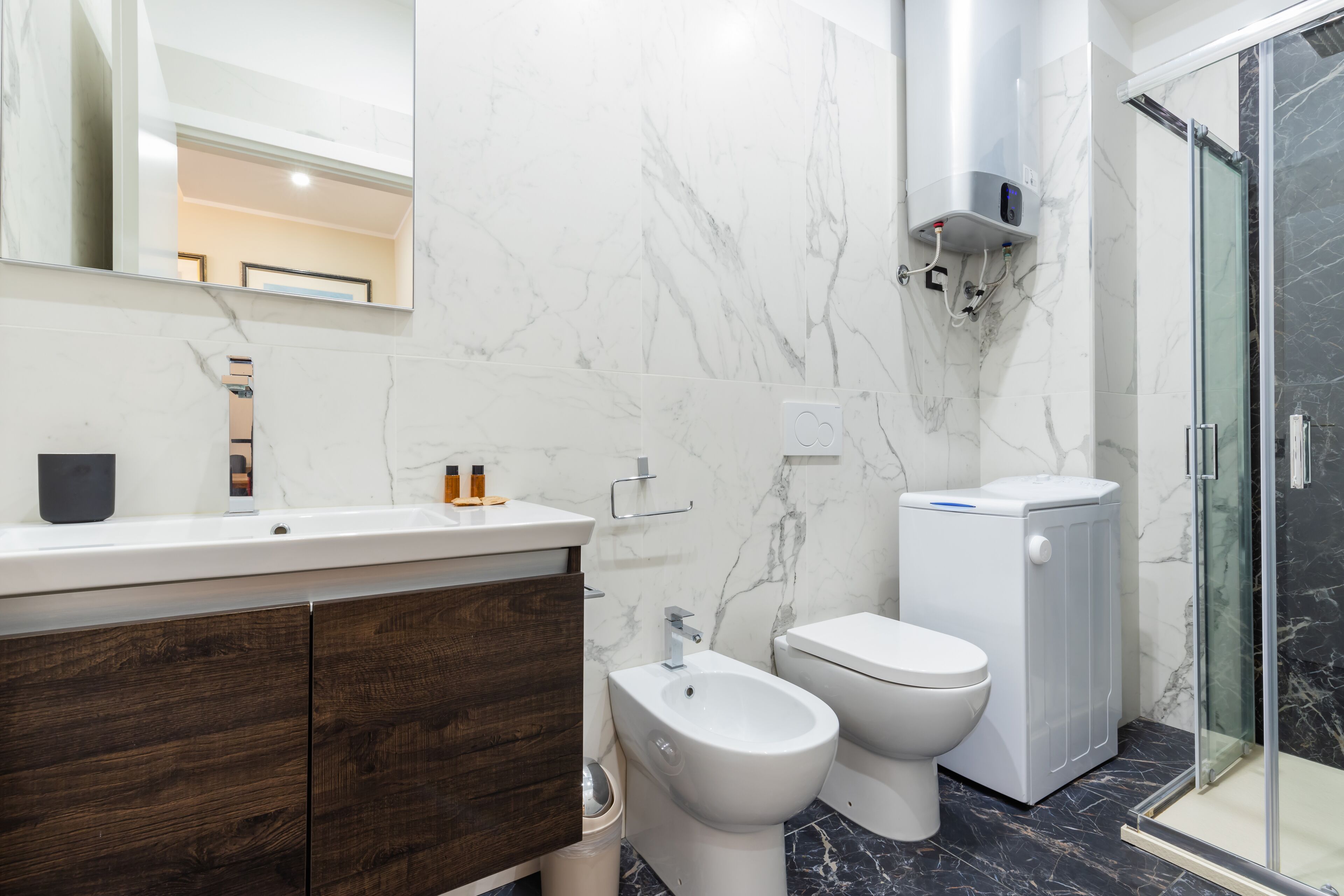 A modern bathroom with a marble wall, a wooden vanity, a bidet, a toilet, and a glass-enclosed shower.