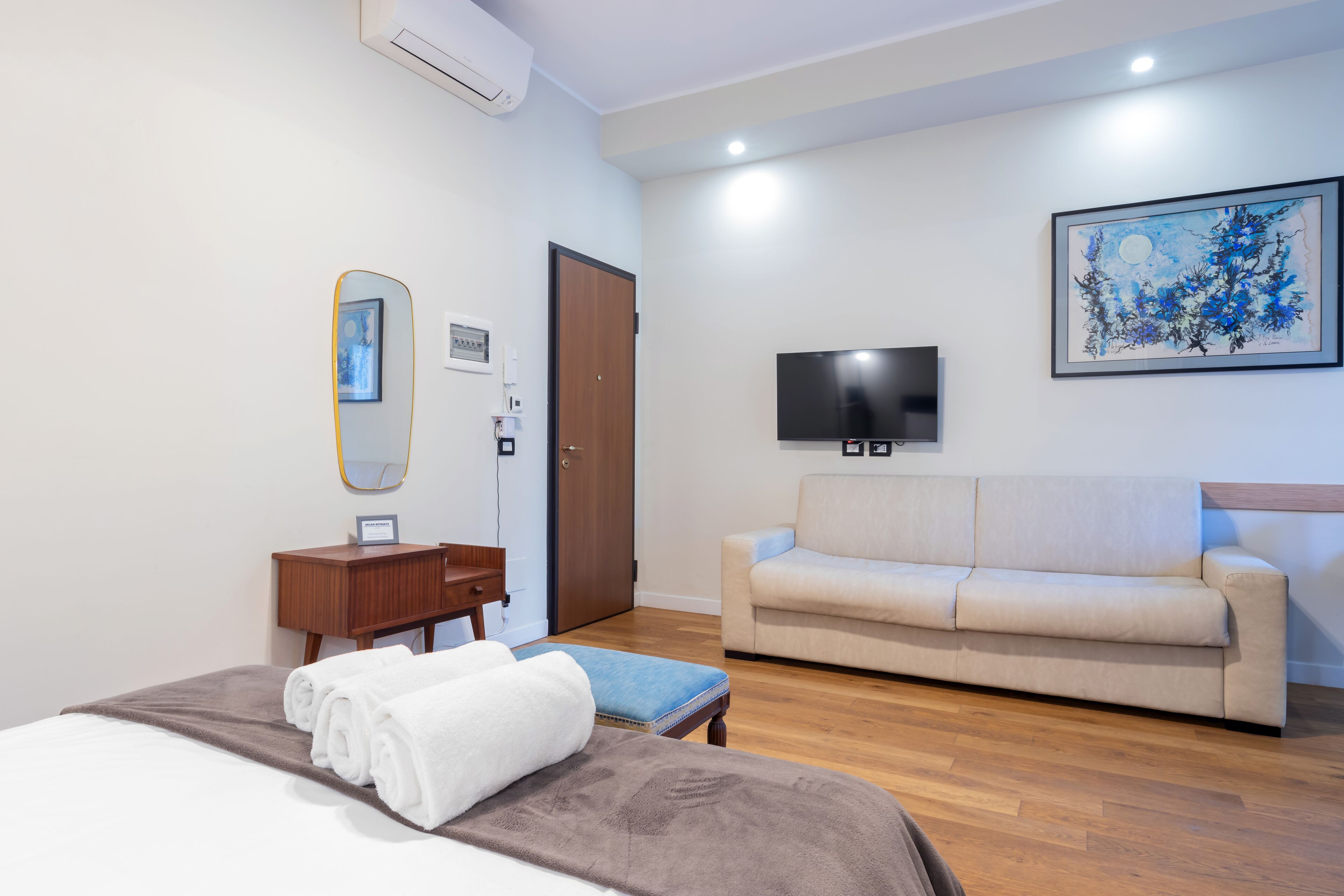 A hotel room with a bed, a wooden nightstand, a small table, a beige sofa, a flat-screen TV, and a framed artwork on the wall.