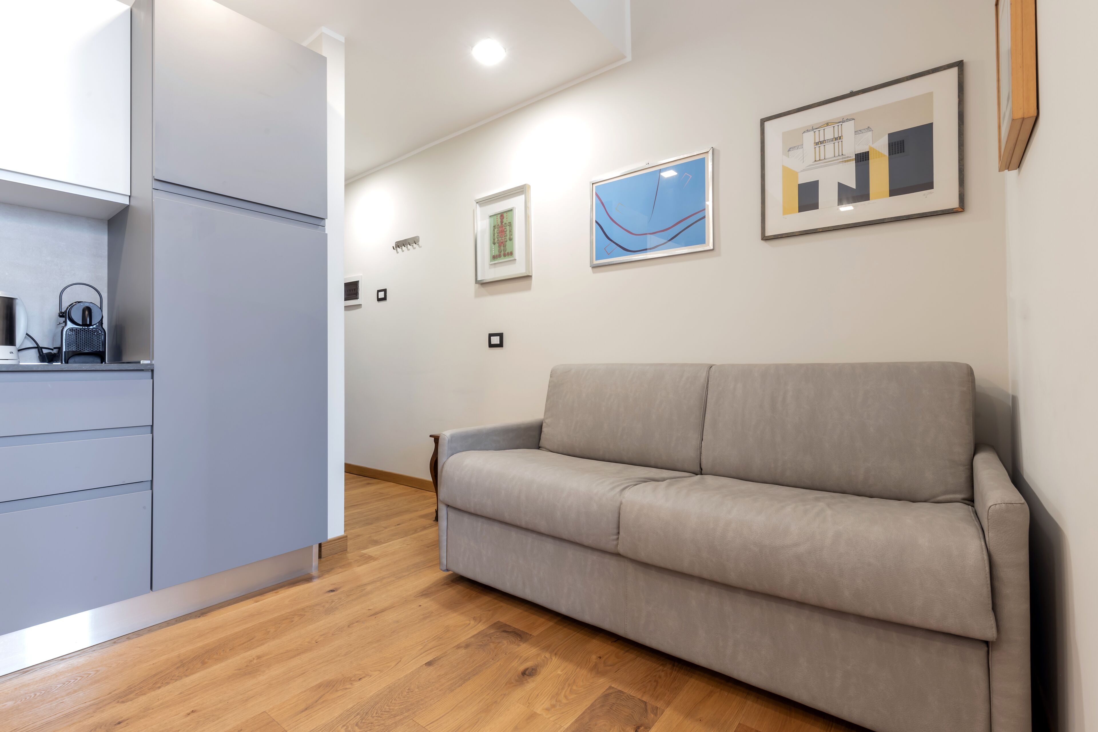 A modern living room with a grey sofa, wooden flooring, and a kitchen area with cabinets and a kettle.