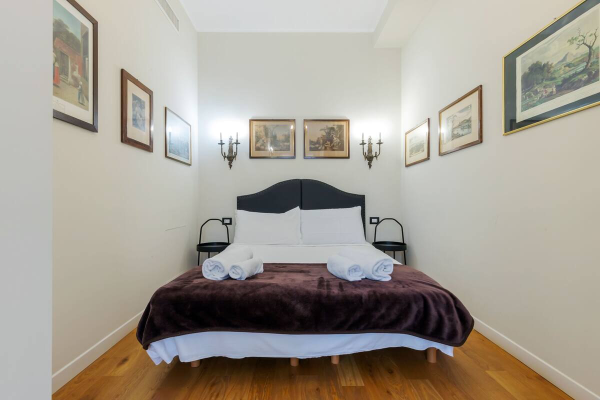 A neatly made bed with a dark brown blanket, white pillows, and two rolled towels. There are framed pictures on the walls and a chandelier.