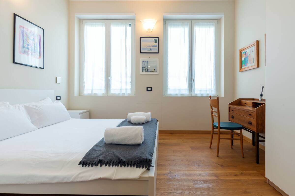 A bedroom with a bed, two windows, a desk, and a chair.