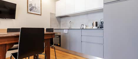A modern kitchen with white cabinets, a wooden dining table with chairs, a built-in oven, and a coffee machine.