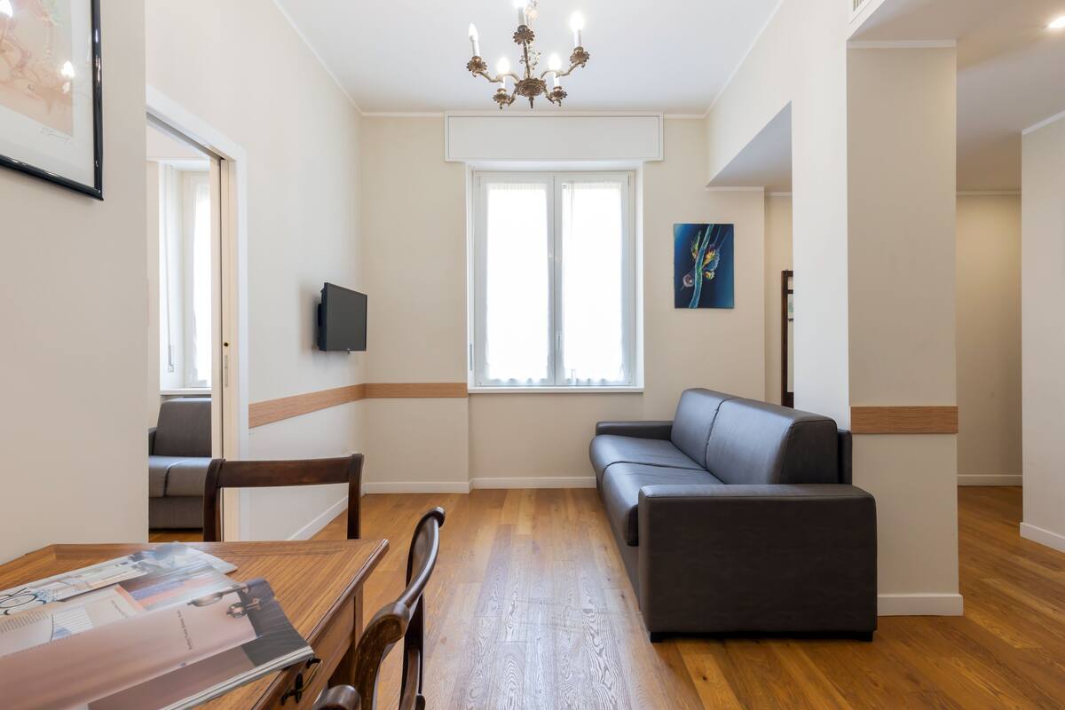 A modern living room with a wooden floor, a grey sofa, a dining table with chairs, a TV, and a chandelier.