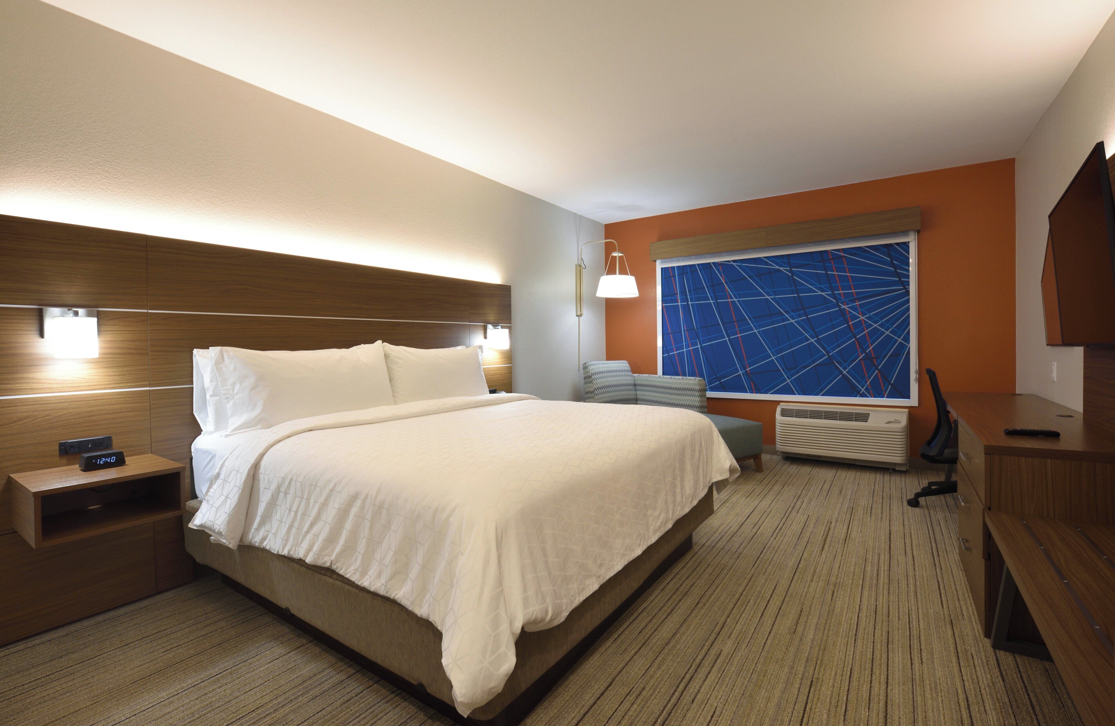 Une chambre d’hôtel avec un grand lit, des tables de chevet, un bureau, une chaise, une télévision et une grande fenêtre donnant sur un paysage aux formes géométriques.