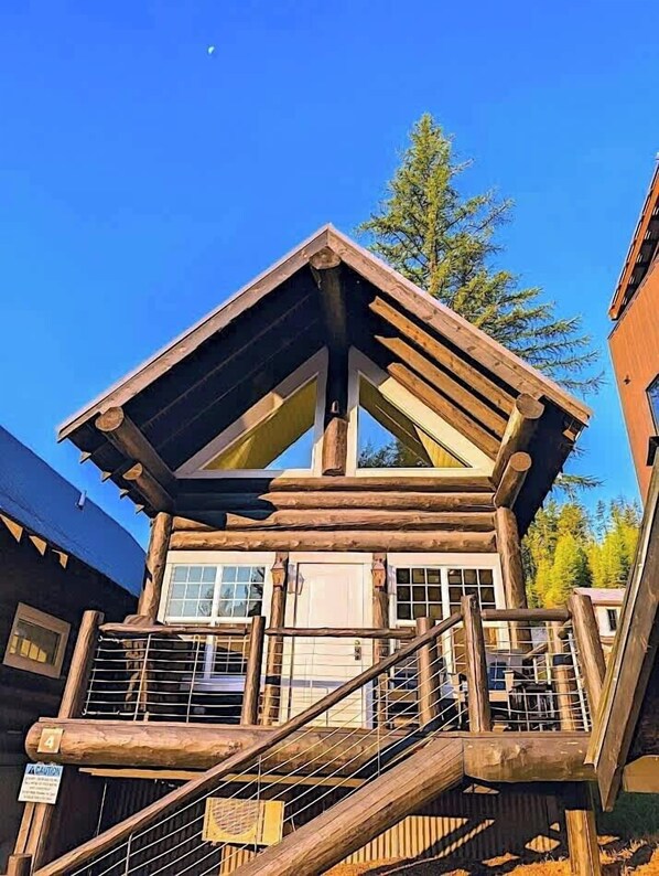 A log cabin with a balcony, a staircase, and a view of the surrounding forest.
