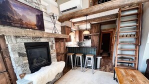 Un interior rústico con chimenea de piedra, vigas de madera y una zona de entrepiso con escalera.