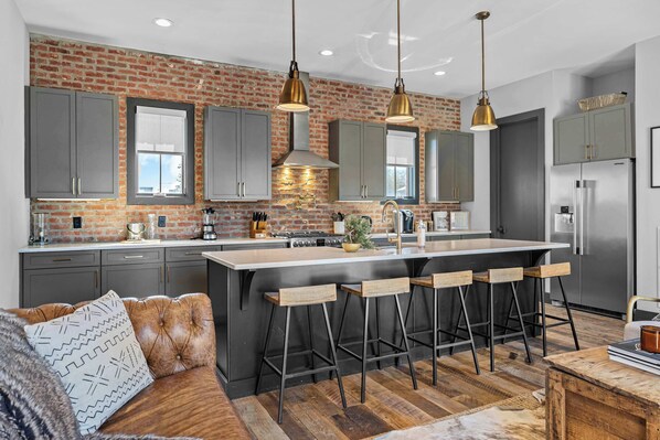 A modern kitchen with a brick wall, grey cabinets, a central island with bar stools, and a stainless steel refrigerator.