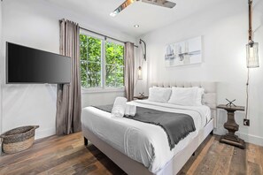 A modern bedroom with a large bed, a flat-screen TV, a window with curtains, and a painting on the wall. - Two Luxury WeHo Homes w/ Rooftops + 6BR Near DT (Nashville)
