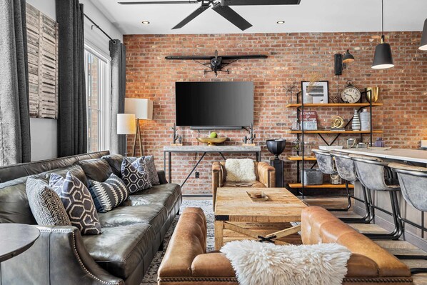 A modern living room with a brick wall, leather furniture, a flat-screen TV, and a dining area with bar stools. - Luxe WeHo Gem | Rooftop Views • DT • Nightlife (Nashville)