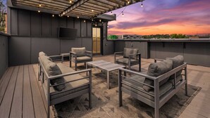Un espacio al aire libre con mobiliario de jardín, una pérgola, sillas con cojines y un televisor de pantalla plana.