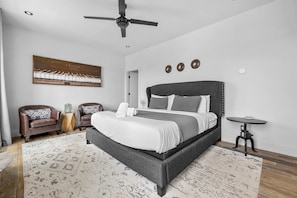 A modern bedroom with a large bed, two leather chairs, a small table, and a ceiling fan.