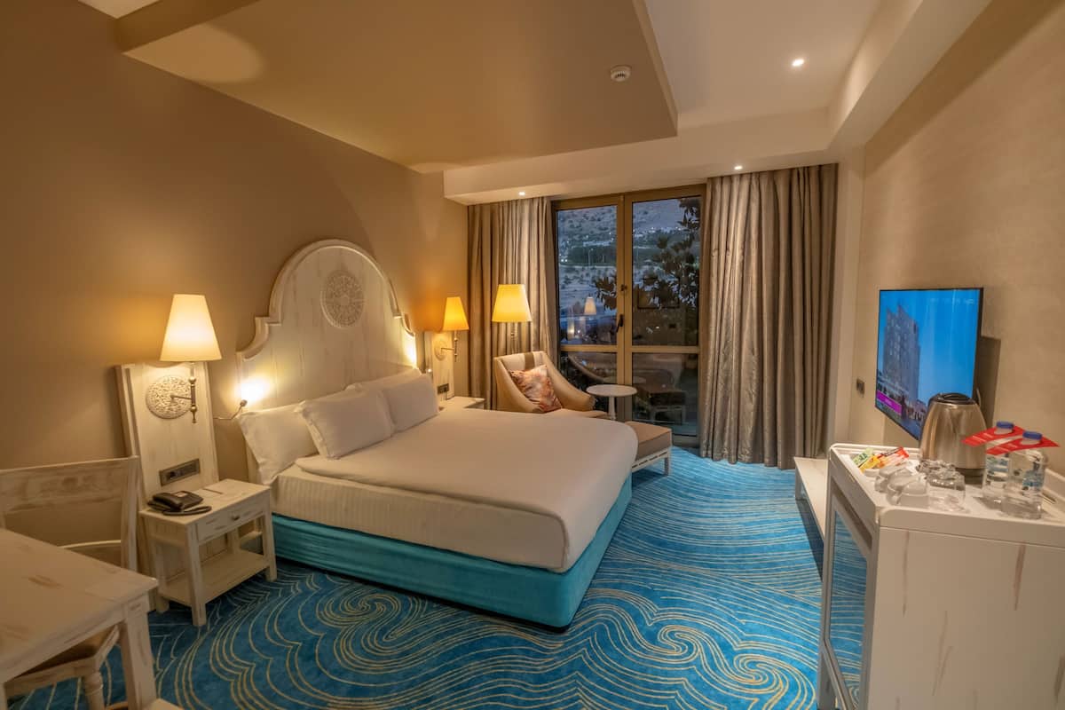 Habitación de hotel con una cama grande, dos lámparas de noche, un televisor, una mesita y vistas al exterior a través de una ventana.