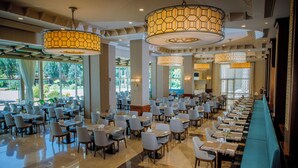 A spacious hotel dining area with multiple tables and chairs, large windows, and decorative lighting fixtures.