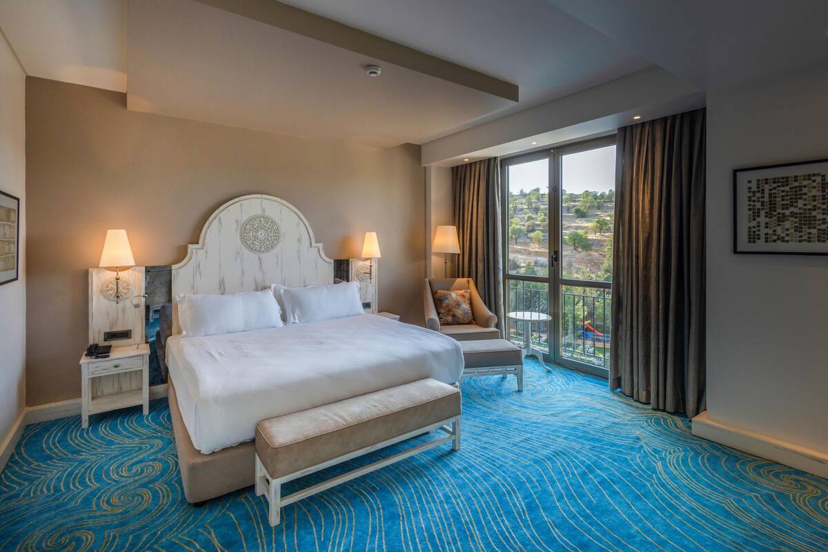 Habitación de hotel con una cama grande, dos mesitas de noche con lámparas, una silla, un balcón con cortinas y vistas al exterior.