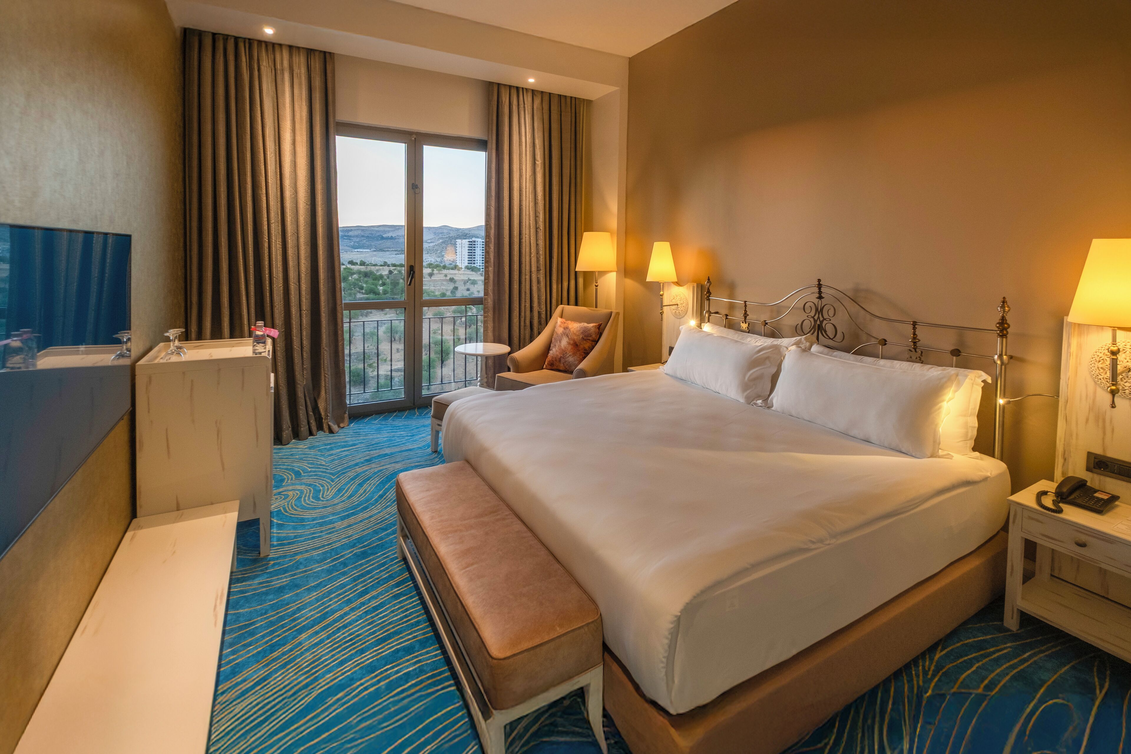 A hotel room with a large bed, a sitting area with a chair and a small table, a desk with a telephone, and a view of the city through a large window.