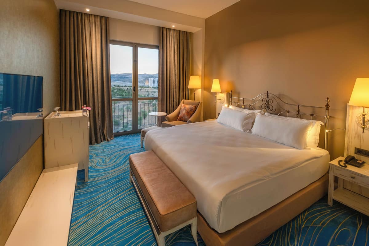 A hotel room with a large bed, a sitting area with a chair and a small table, a desk with a telephone, and a view of the city through a large window.