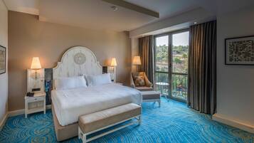 Habitación de hotel con una cama grande, dos mesitas de noche con lámparas, una silla, un balcón con vistas y una alfombra azul estampada.