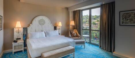 Habitación de hotel con una cama grande, dos mesitas de noche, una silla y vistas a un paisaje a través de un ventanal.