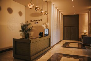 Ein Empfangsschalter mit einer an der Wand befestigten Schilder, auf dem 'ELEGANCE SPA' steht, einer Topfpflanze und einem Bildschirm, der ein Logo zeigt.