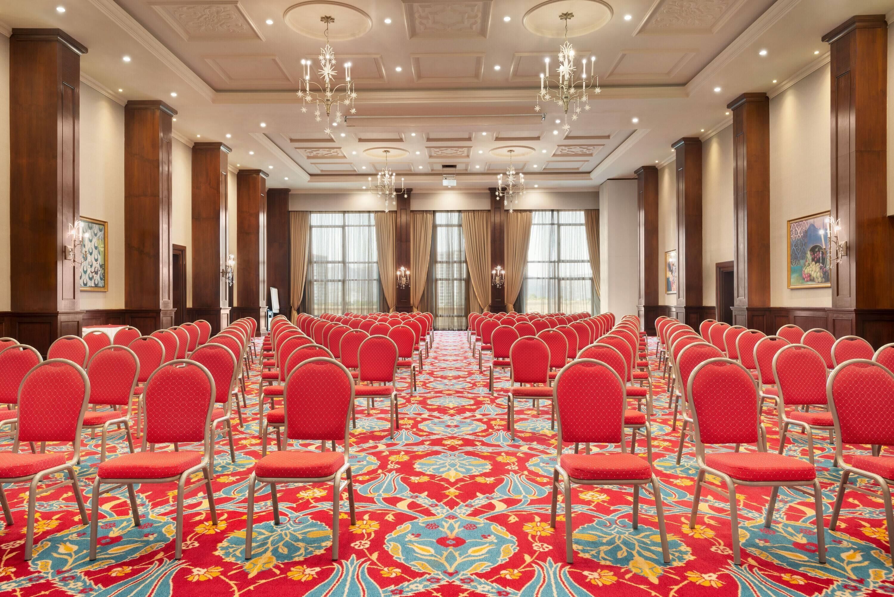 Une salle de conférence avec des rangées de chaises rouges, une moquette à motifs, des lustres et de grandes fenêtres avec des rideaux.
