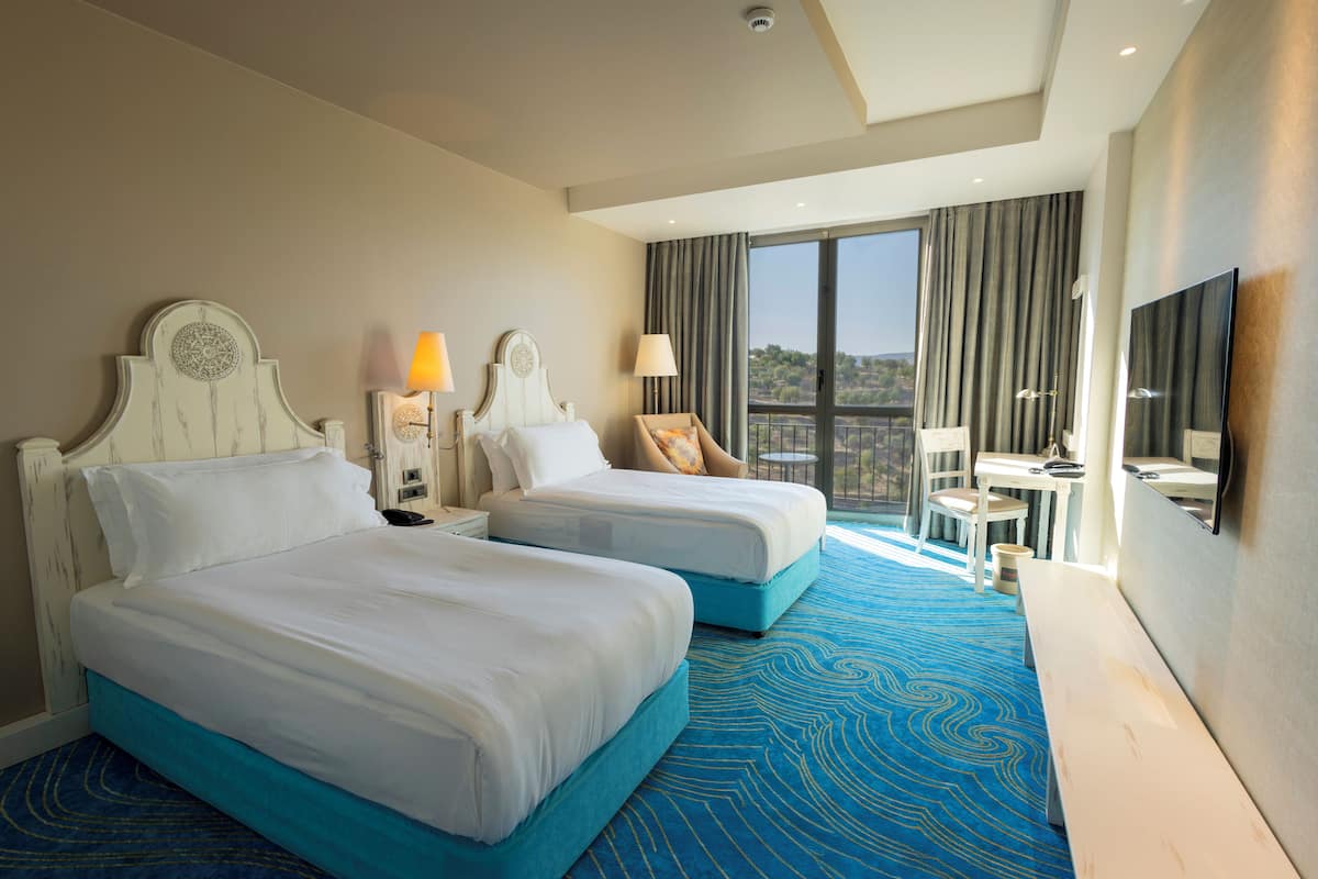 Habitación de hotel con dos camas, un escritorio, una silla, un televisor y un balcón con vistas.