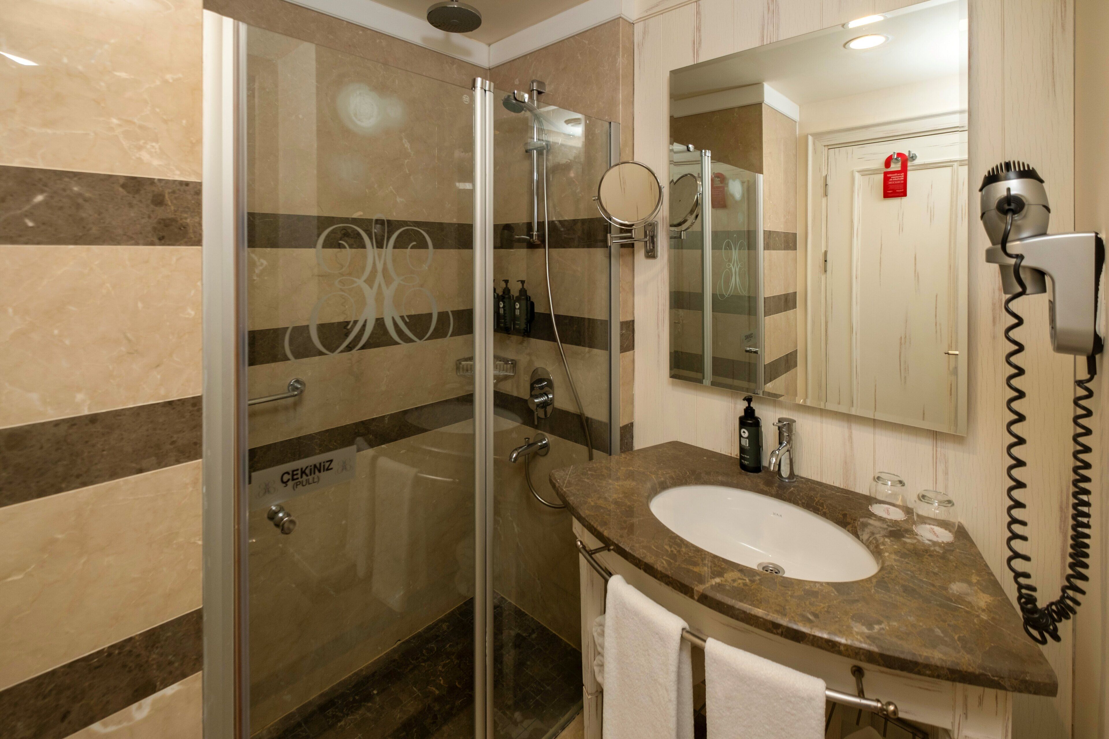 Un baño moderno con ducha envuelta en cristal, lavamanos con encimera de mármol y un secador de pelo montado en la pared.