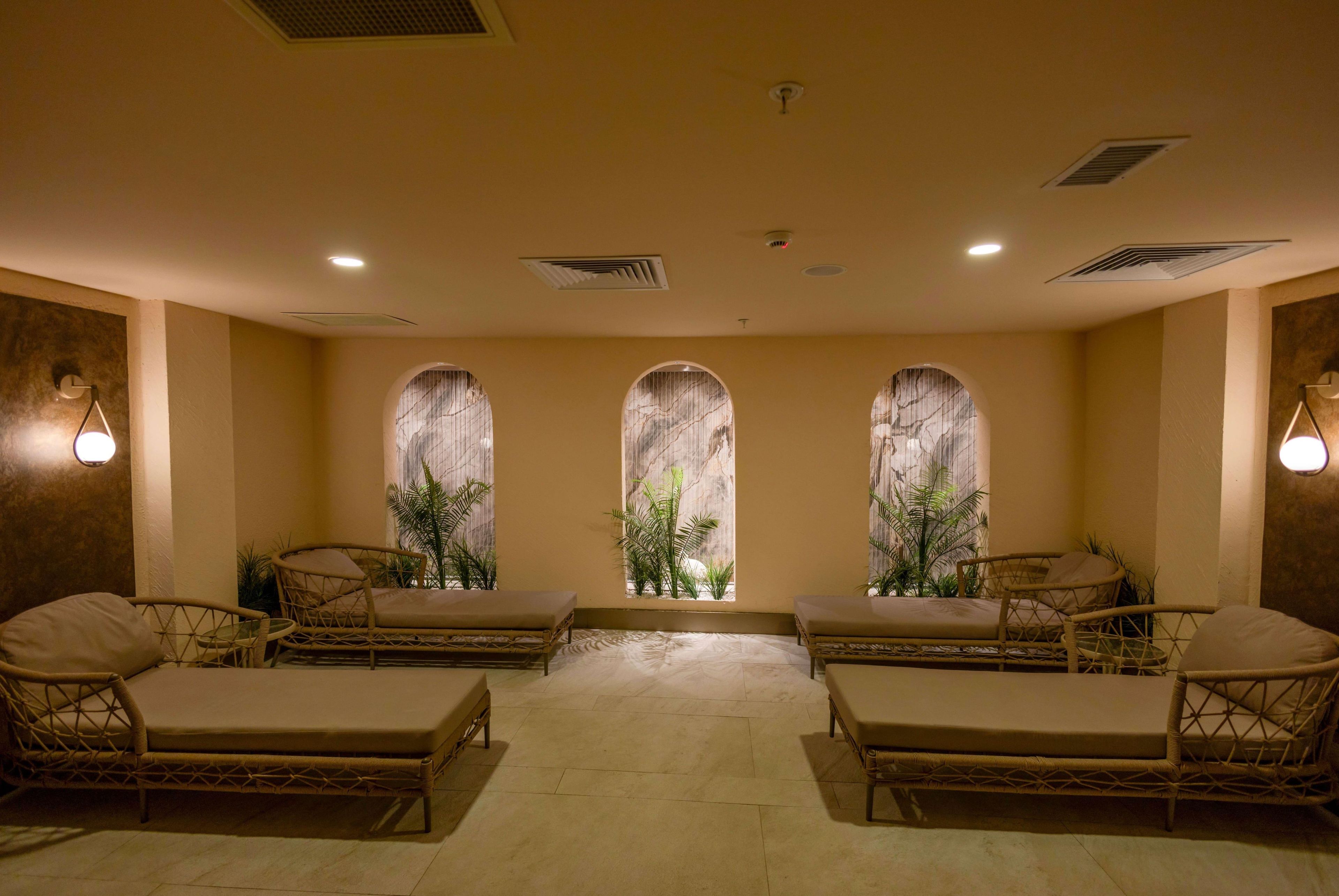 Una habitación con cuatro sillas de mimbre, una mesita pequeña y tres ventanas arqueadas con plantas.