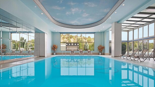 A large indoor swimming pool with clear glass walls, a view of trees outside, and a ceiling with a decorative pattern.
