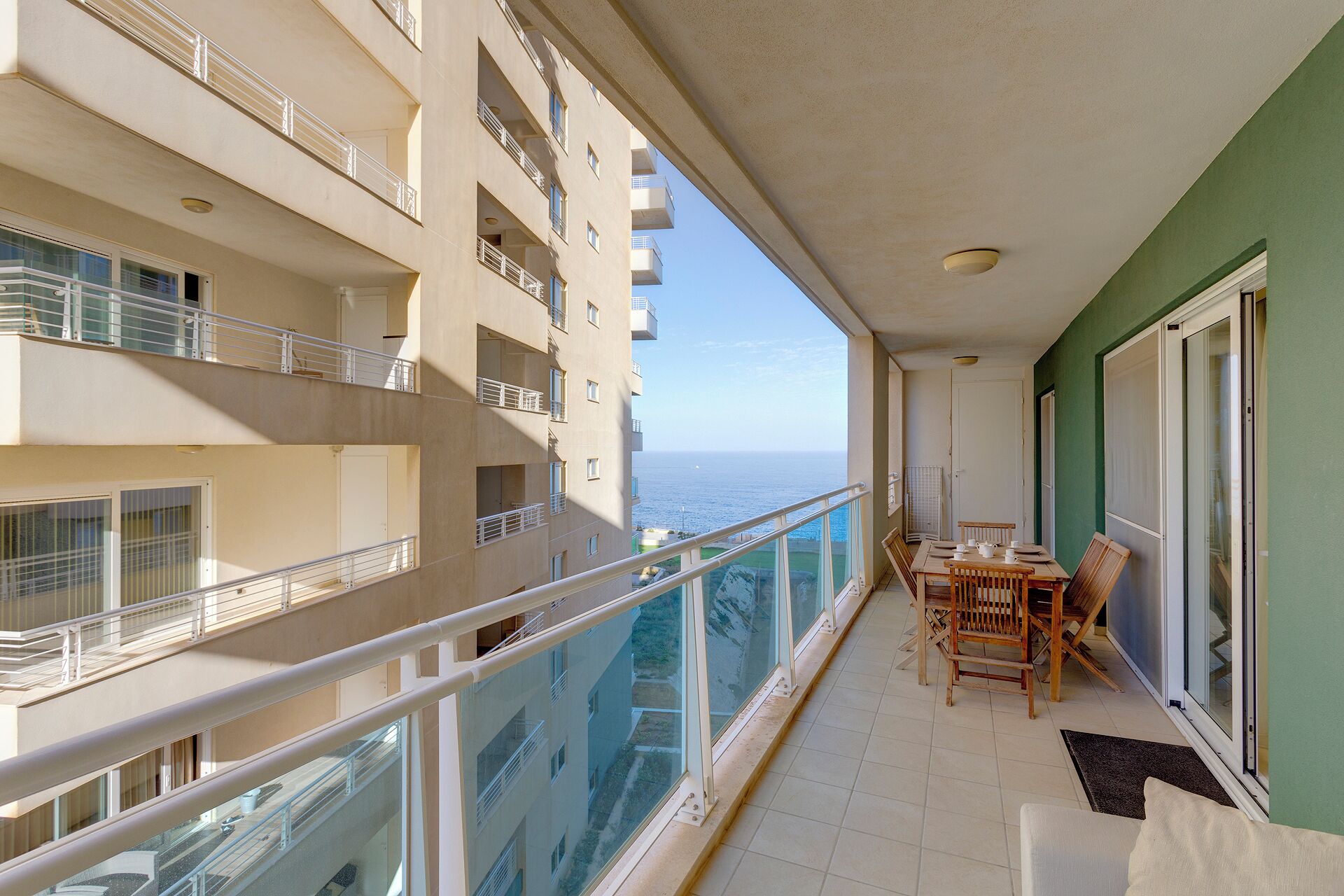 A balcony with a dining table and chairs, overlooking a building with multiple balconies and a view of the sea.
