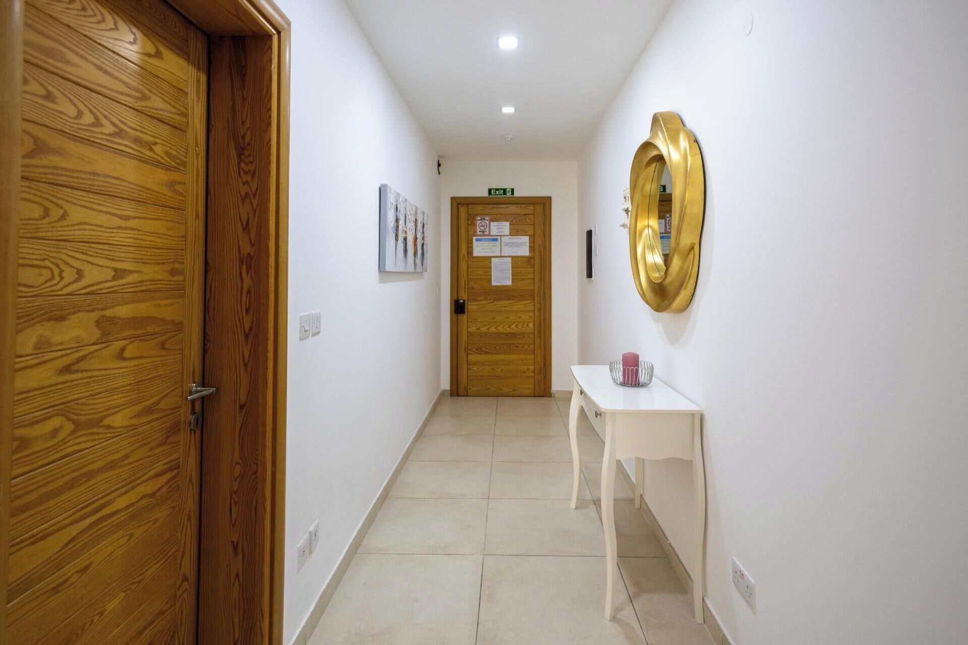 A hallway with a wooden door, a white wall with a gold mirror, and a white table with a pink object.