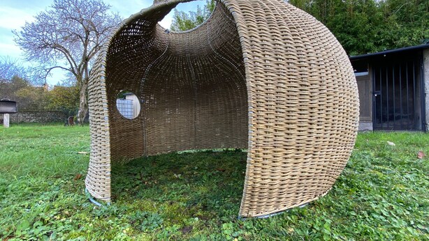 Une structure en osier, dotée d’une ouverture circulaire, installée sur une pelouse avec des arbres et un bâtiment en arrière-plan.