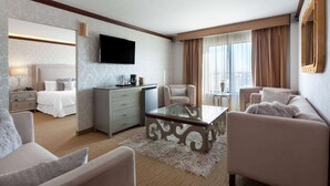 Quarto de hotel com cama, área de estar com sofá e poltronas, mesa de centro, televisão de tela plana na parede, mesinha de cabeceira com abajur e uma janela grande com cortinas.
