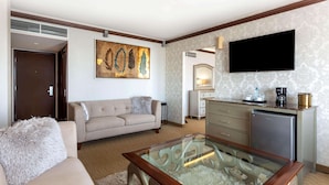 Quarto de hotel com sofá bege, mesa de centro de vidro, televisão de tela plana, frigobar e um quadro na parede.