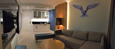 A modern living room with a sofa, ottoman, and round coffee table. A wall-mounted TV and a lamp are visible.