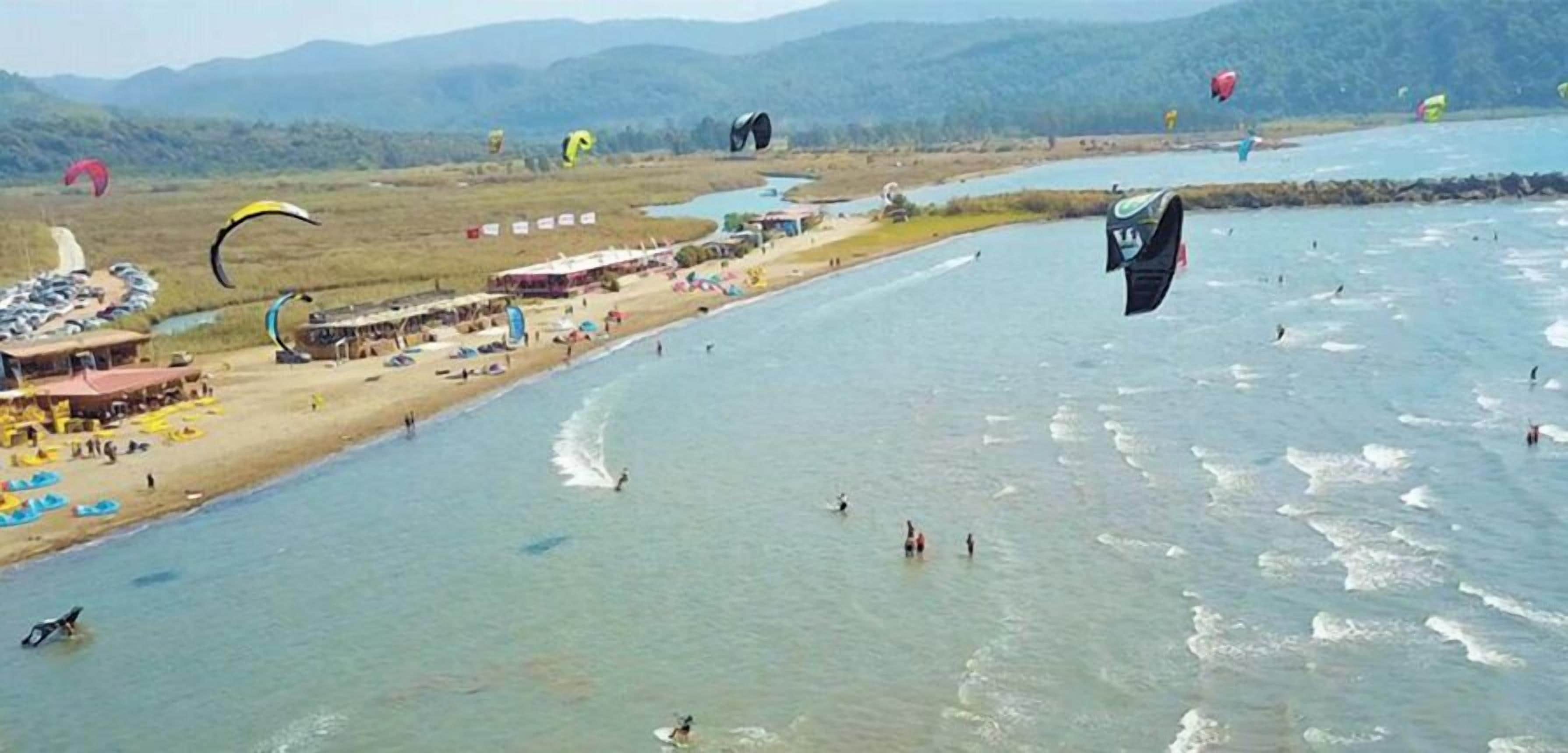 Una playa con actividad de parasailing, un estacionamiento y un río.