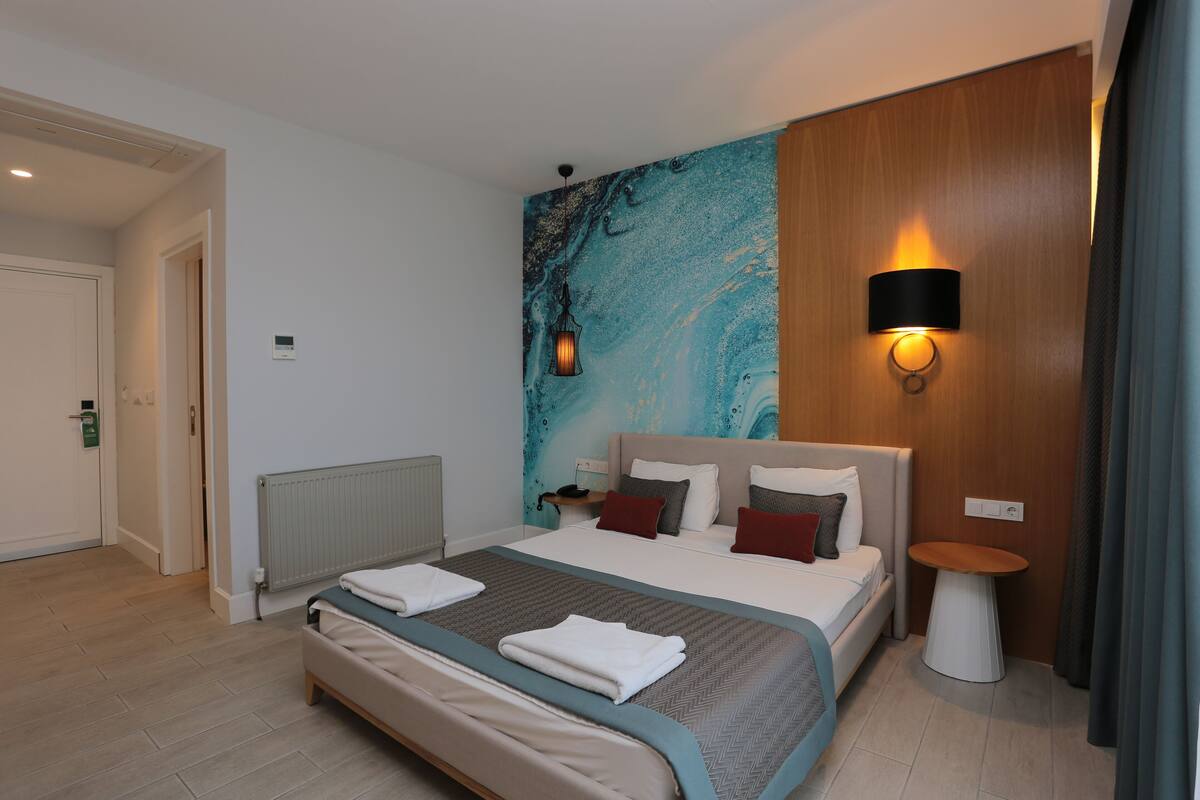 A modern hotel room with a large bed, a wall-mounted lamp, and a small side table.