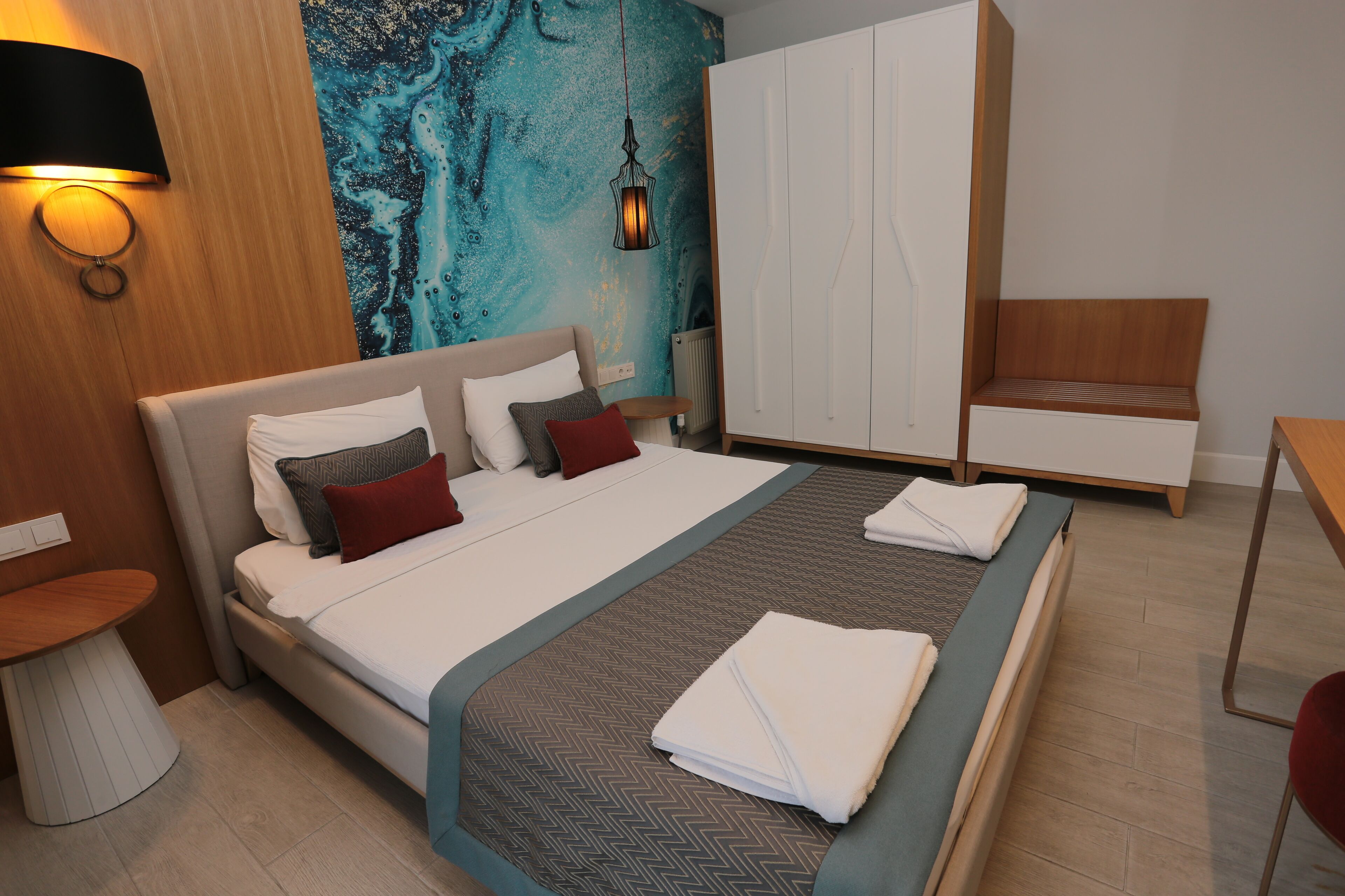 A modern hotel room with a large bed, a wooden side table, a small round table, a wardrobe, and a bench.