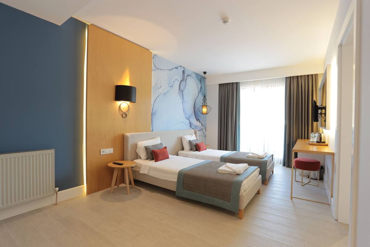 A hotel room with two beds, a desk, a chair, a wall-mounted lamp, and a window with curtains.