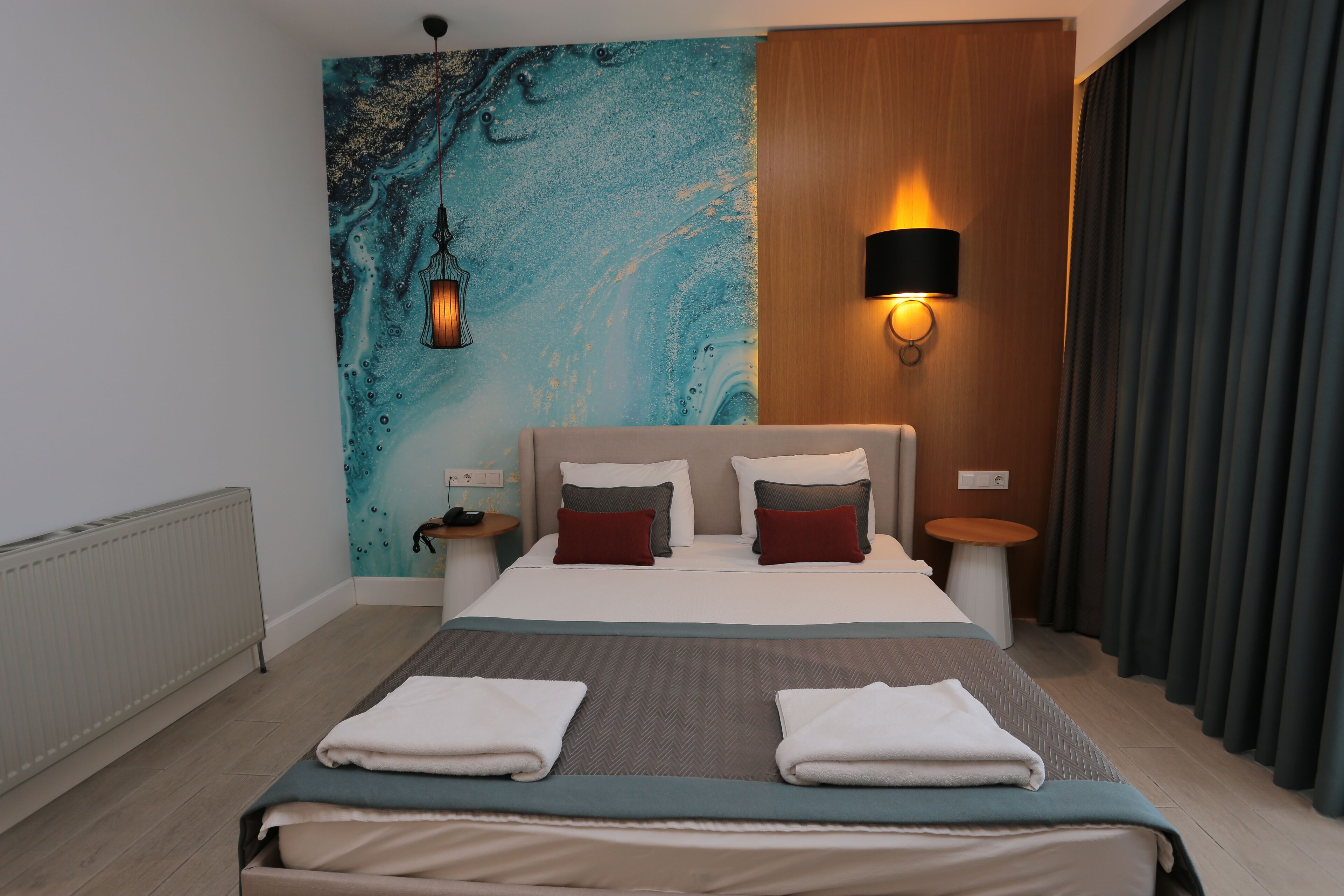 A hotel room with a large bed, a wall-mounted lamp, a small table, and a view of the outside through curtains.