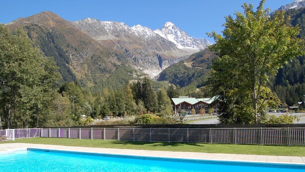 A swimming pool with a mountain backdrop, a fenced area, and a lodge with a green roof. - Résidence Grands Montets 211 Walk to Ski Lift | Argentière Chamonix, Argentière (Chamonix), France (Argentière (Chamonix))