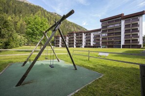 A playground with swings and a bench, surrounded by a grassy area and a multi-story building with balconies.
